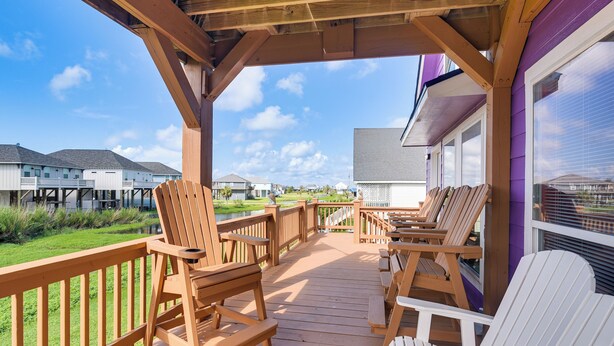 Una terraza de madera con mobiliario exterior, un espacio cubierto y vistas a casas al fondo.