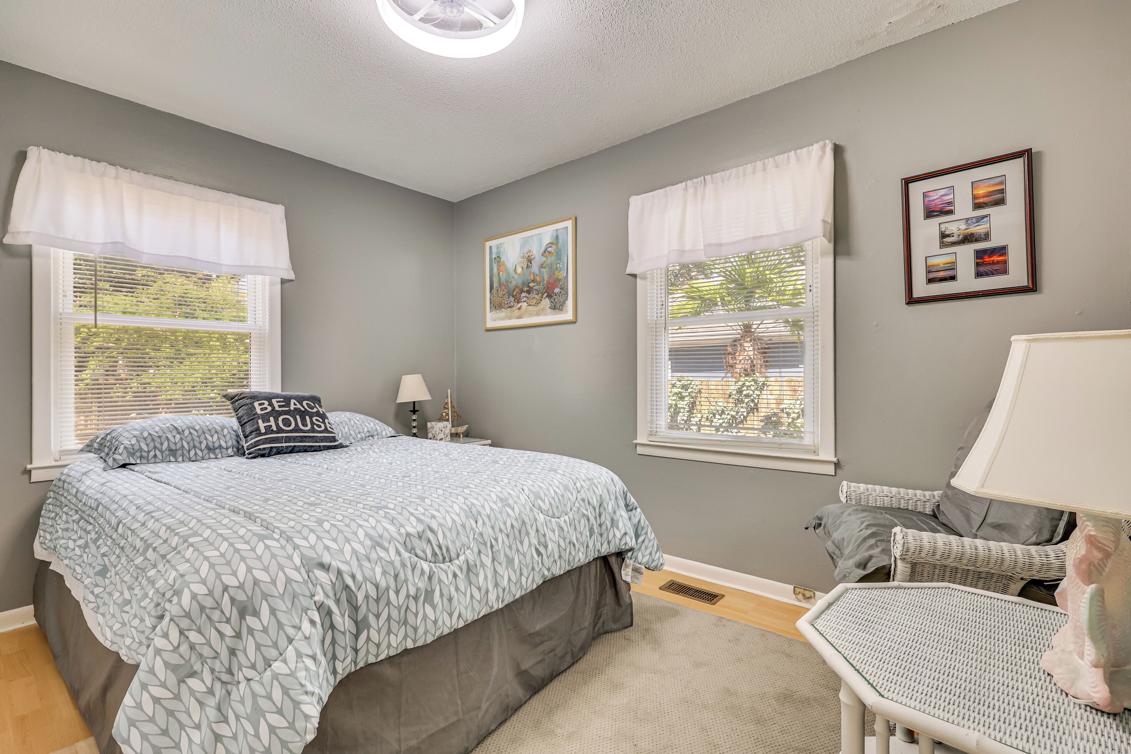 A bedroom with a bed, two windows with white valances, a bedside table with a lamp, a chair, and wall art.