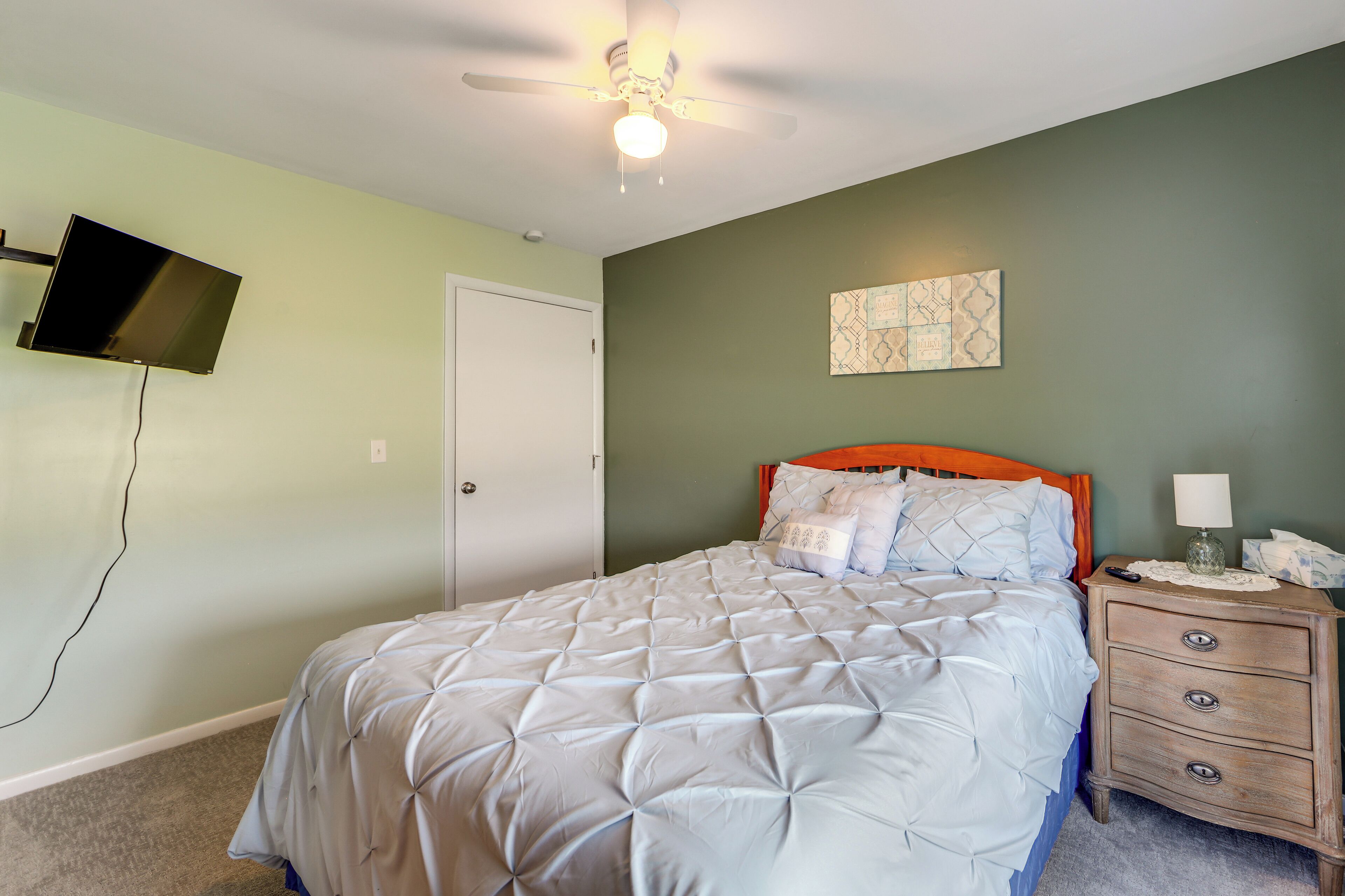 A bedroom with a bed, a nightstand, a wall-mounted TV, a ceiling fan, and a door.