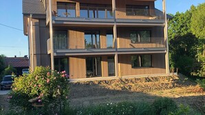Un edificio residencial de varios pisos con balcones, un jardín con flores y un coche estacionado al fondo.