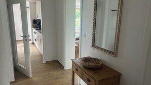 Un pasillo con piso de madera, una consola de madera con cajones, un espejo grande con marco decorativo, y una puerta que da a una zona de cocina.