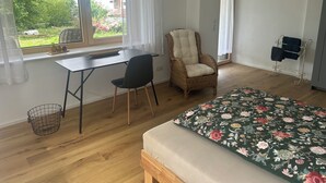 Una habitación con una cama, un escritorio, una silla, un ventanal con cortinas, una silla de mimbre y un estante metálico con toallas.