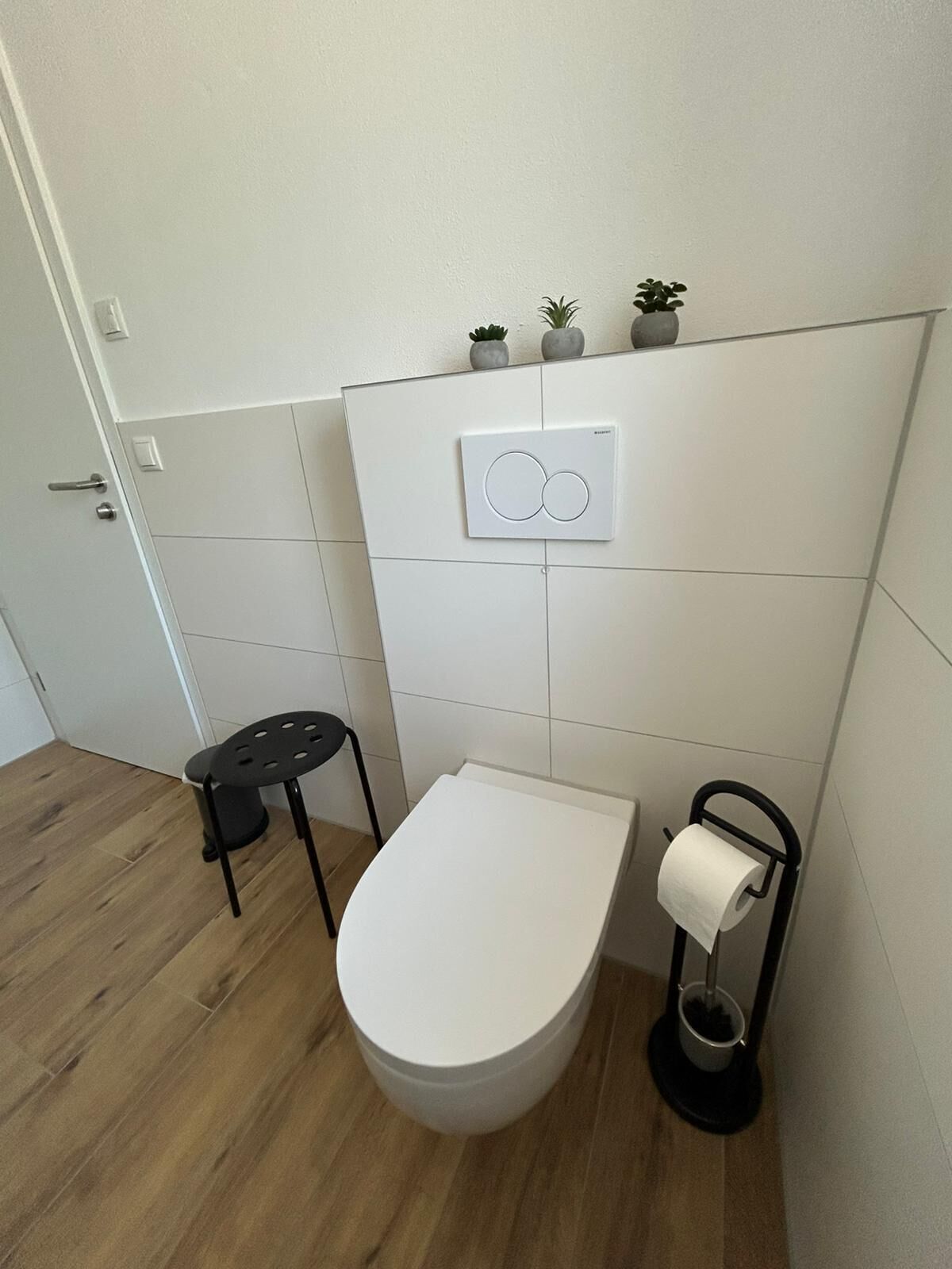 A modern bathroom with a toilet, a small stool, a toilet paper holder, and a wall-mounted light switch.