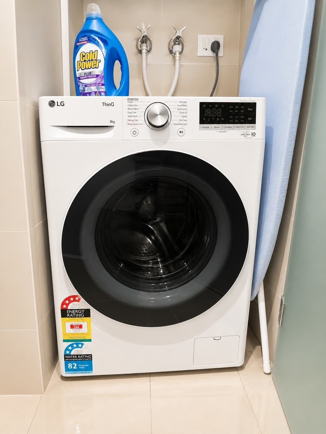 Een voorlader wasmachine van LG met een digitaal display, energielabels en een waterverbruiklabel, en een blauwe fles wasmiddel ernaast.