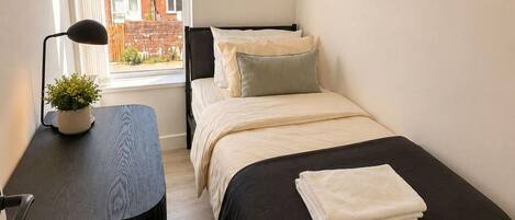 A small, tidy bedroom with a single bed, a bedside table, a lamp, and a window with a view of a brick building.