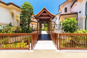 A covered walkway with wooden railings, flanked by lush greenery and buildings with Mediterranean-style architecture. - Casa Luna 4: Gated Luxury Near 7Mile Beach w/Pool, Heated Spa, and Private Beach Cove (Grand Cayman)