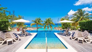 A wooden deck with a pool, lounge chairs, and umbrellas. - Casa Luna 4: Gated Luxury Near 7Mile Beach w/Pool, Heated Spa, and Private Beach Cove (Grand Cayman)