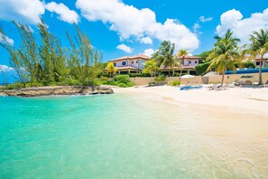 A beachfront property with a pool, palm trees, and a clear blue sky. - Casa Luna 4: Gated Luxury Near 7Mile Beach w/Pool, Heated Spa, and Private Beach Cove (Grand Cayman)