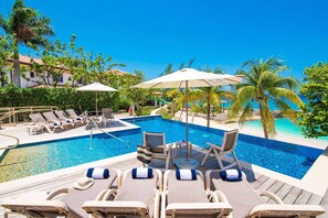 A pool area with lounge chairs and umbrellas, surrounded by palm trees and a clear blue sky. - Casa Luna 4: Gated Luxury Near 7Mile Beach w/Pool, Heated Spa, and Private Beach Cove (Grand Cayman)