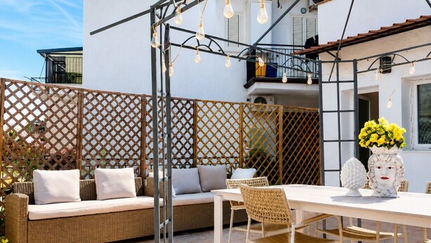 Eine Terrasse im Freien mit einem Korbsessel, einem Esstisch, Stühlen und einer dekorativen Vase mit Blumen.