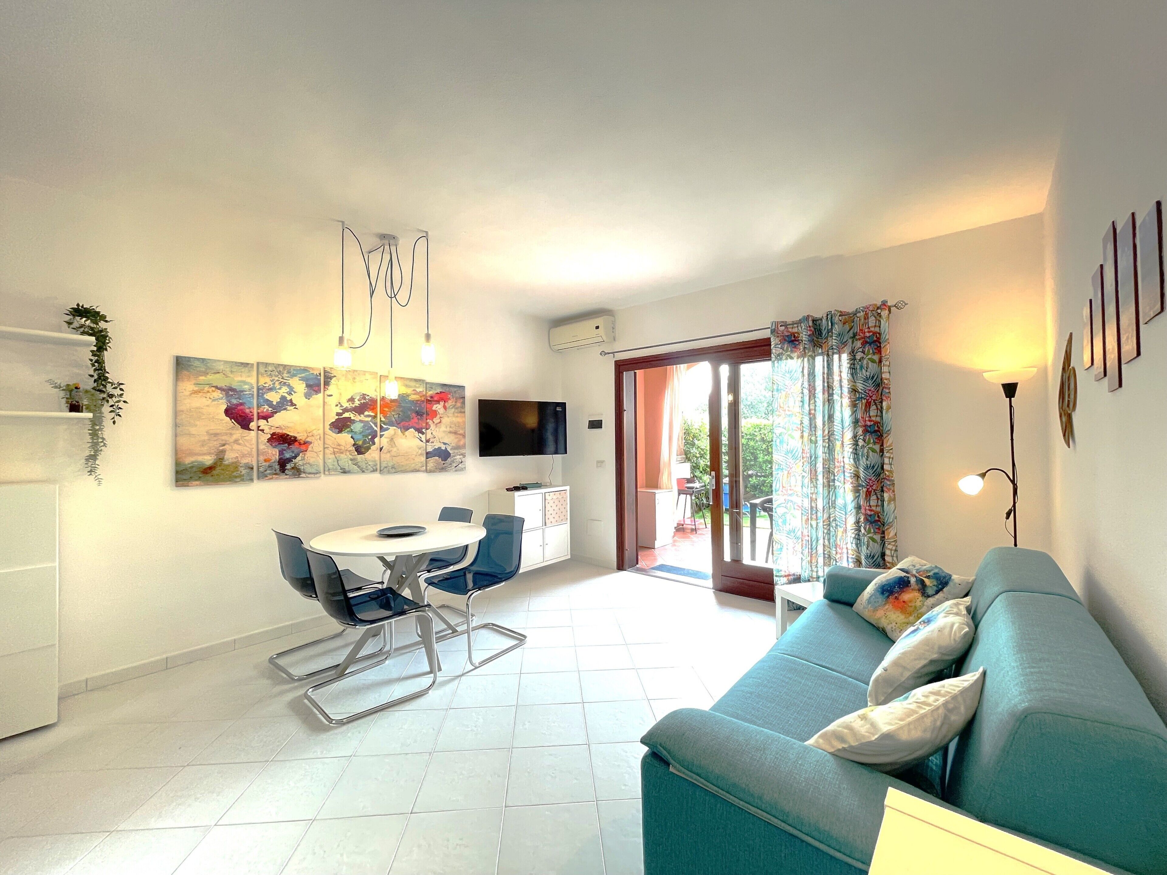 A modern living room with a sofa, a dining table with chairs, a TV mounted on the wall, and a sliding glass door leading to an outdoor area.
