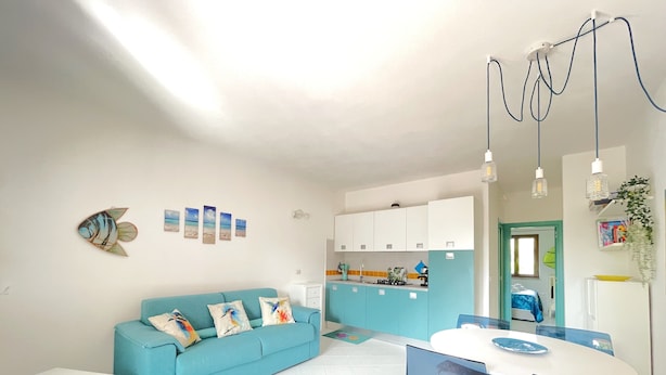Een knus woongedeelte met een blauwe bank, een witte eettafel met stoelen, een kitchenette en een kleine keukenhoek.