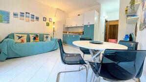Een ruimtelijk ingerichte leefruimte met kitchenette, een eethoek met ronde tafel en een bank met kleurrijke kussens.