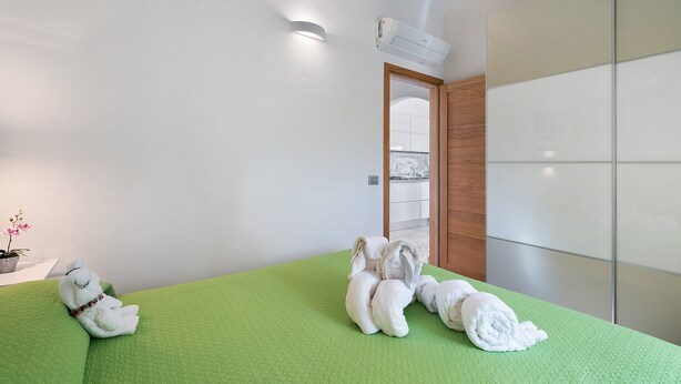 Hotellihuone, jossa on vihreä, kuvioitu sängynpeite, seinälle asennettu ilmastointilaite ja puuovi, joka johtaa parvekkeelle.