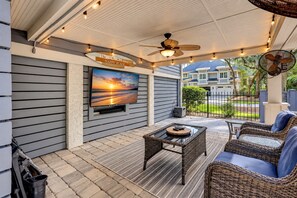 Ein überdachter Patio-Bereich mit an der Wand montiertem Fernseher, Korbmöbeln und einem Deckenventilator.