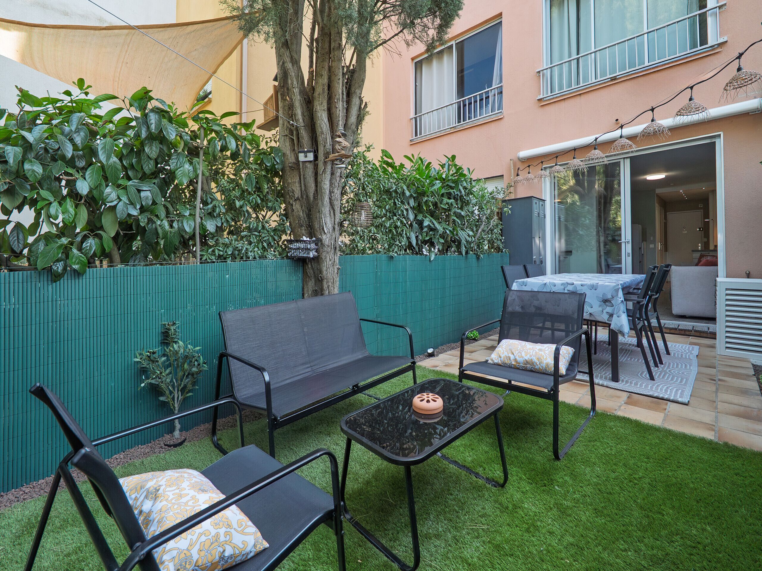 Une terrasse avec un canapé, des chaises et une table, entourée d’une clôture verte et d’arbres.