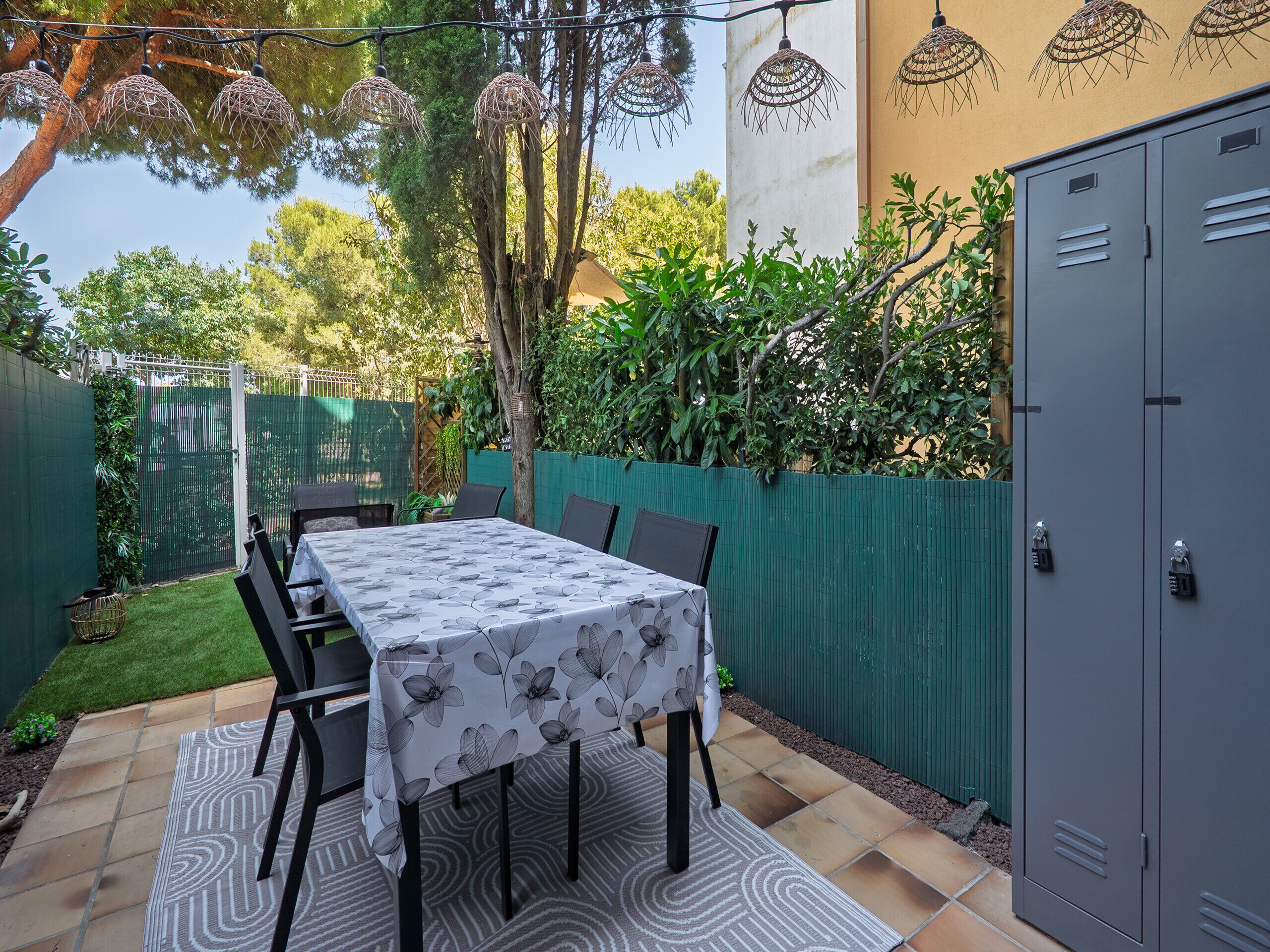 Un patio con mesa lista para comer, rodeado por una cerca verde y árboles.