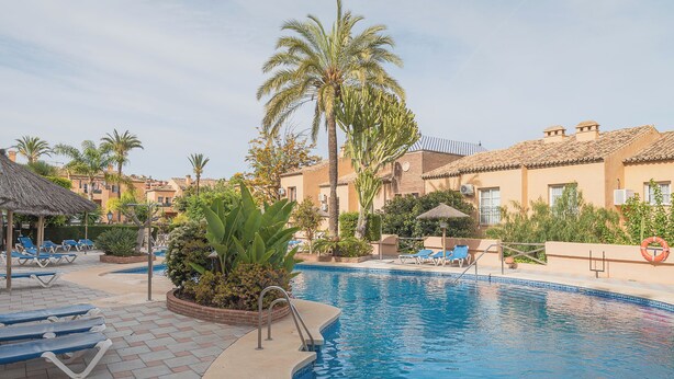 Zona de piscina con sillas de descanso, palmeras y un edificio con tejado de azulejos.