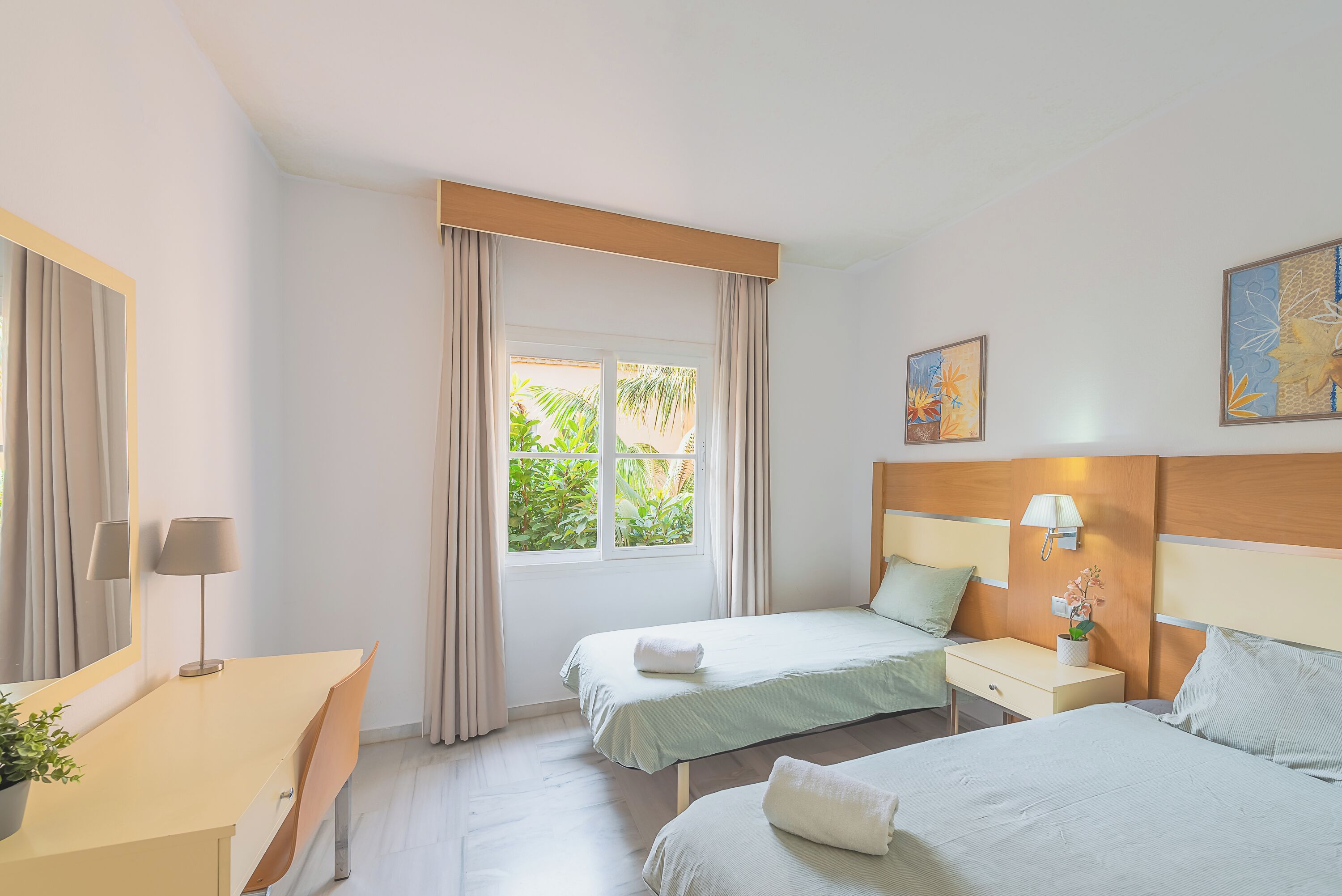 Habitación de hotel con dos camas, un escritorio, una silla, ventana con cortinas y una lámpara de pared.