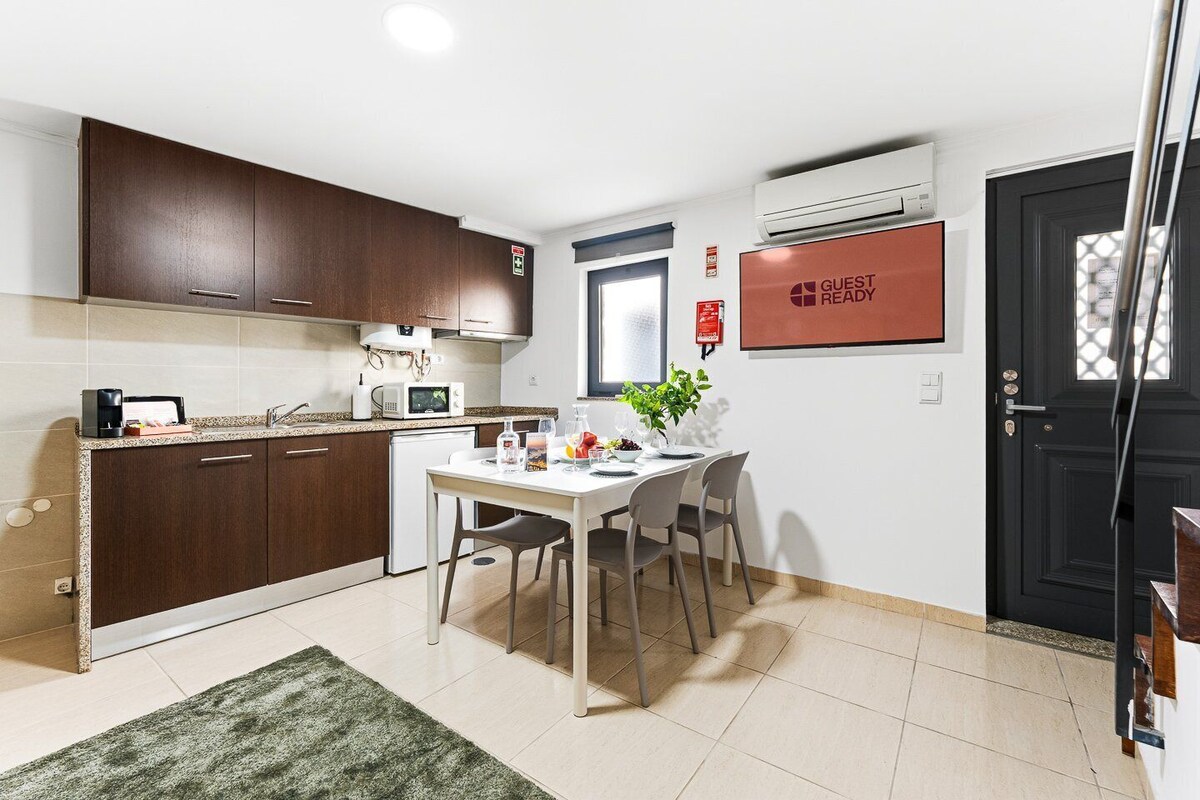 A compact kitchen with dark cabinets, a white countertop, a dining table with chairs, a small refrigerator, and a 'GUEST READY' sign.