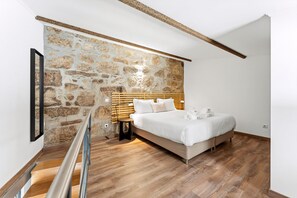 Un dormitorio con pared de piedra, una cama con ropa blanca, una mesita de noche pequeña y suelo de madera.