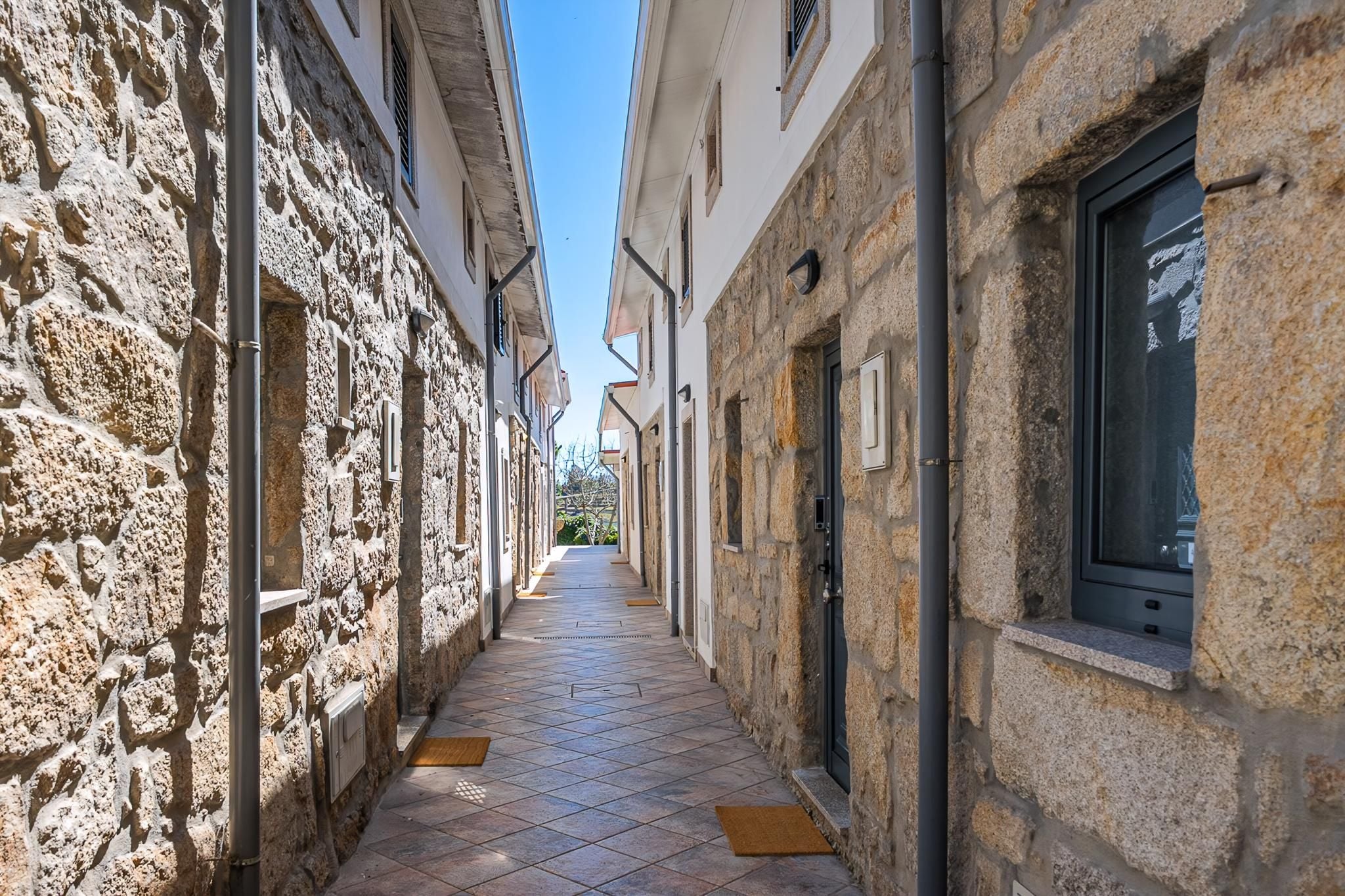 A narrow stone corridor with tiled flooring, flanked by buildings with stone walls and windows.