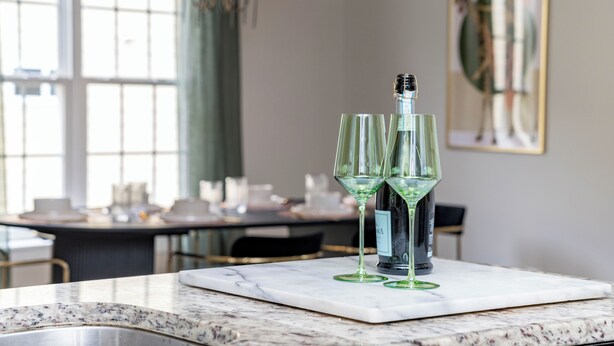 Een marmeren aanrecht met twee groene wijnglazen en een fles, een eettafel op de achtergrond, en een ingelijst schilderij aan de muur.