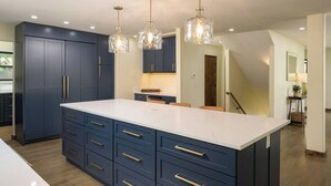 Een moderne keuken met donkerblauwe kastjes, een groot eiland met lades en drie hanglampen.