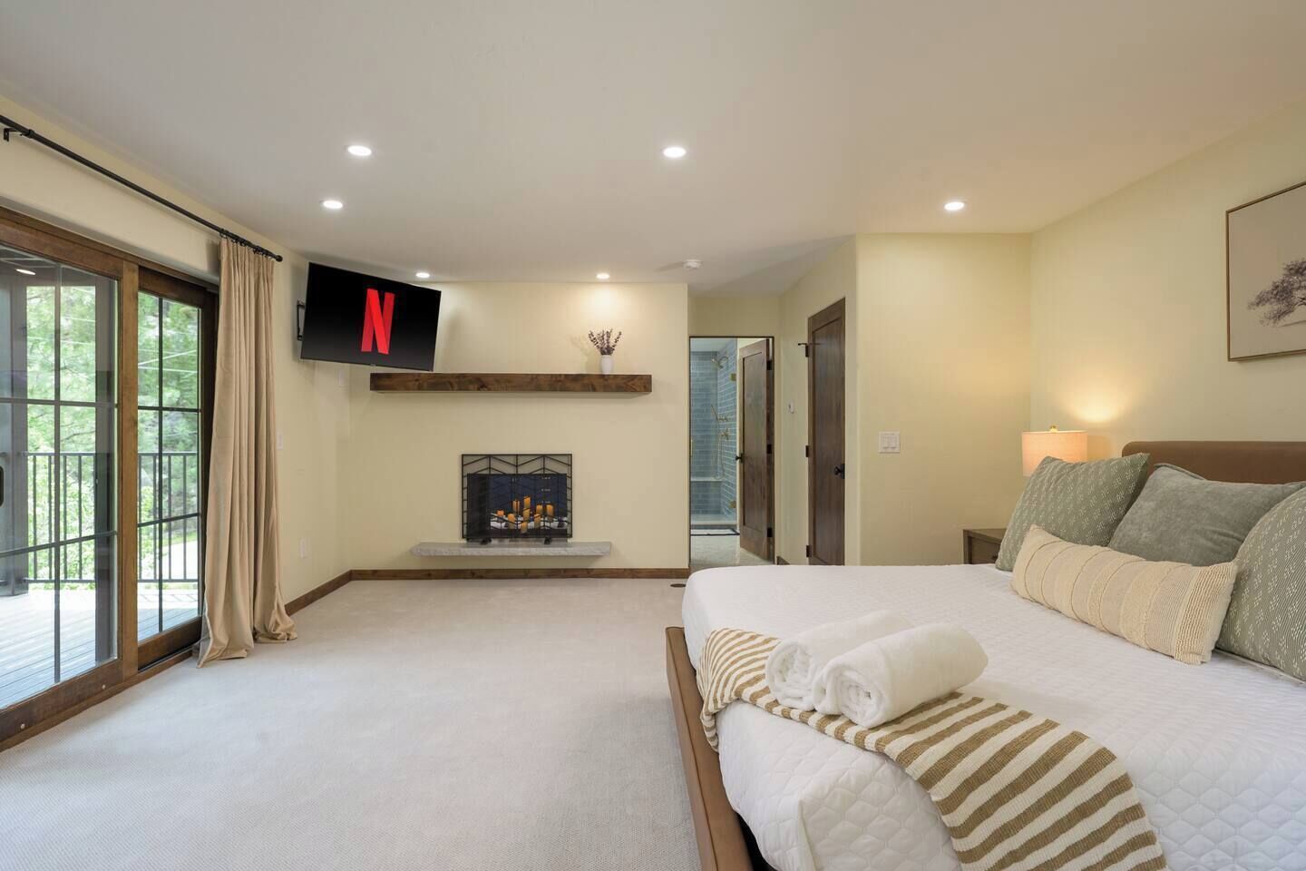 A bedroom with a bed, a TV mounted on the wall, a fireplace, and a large window.