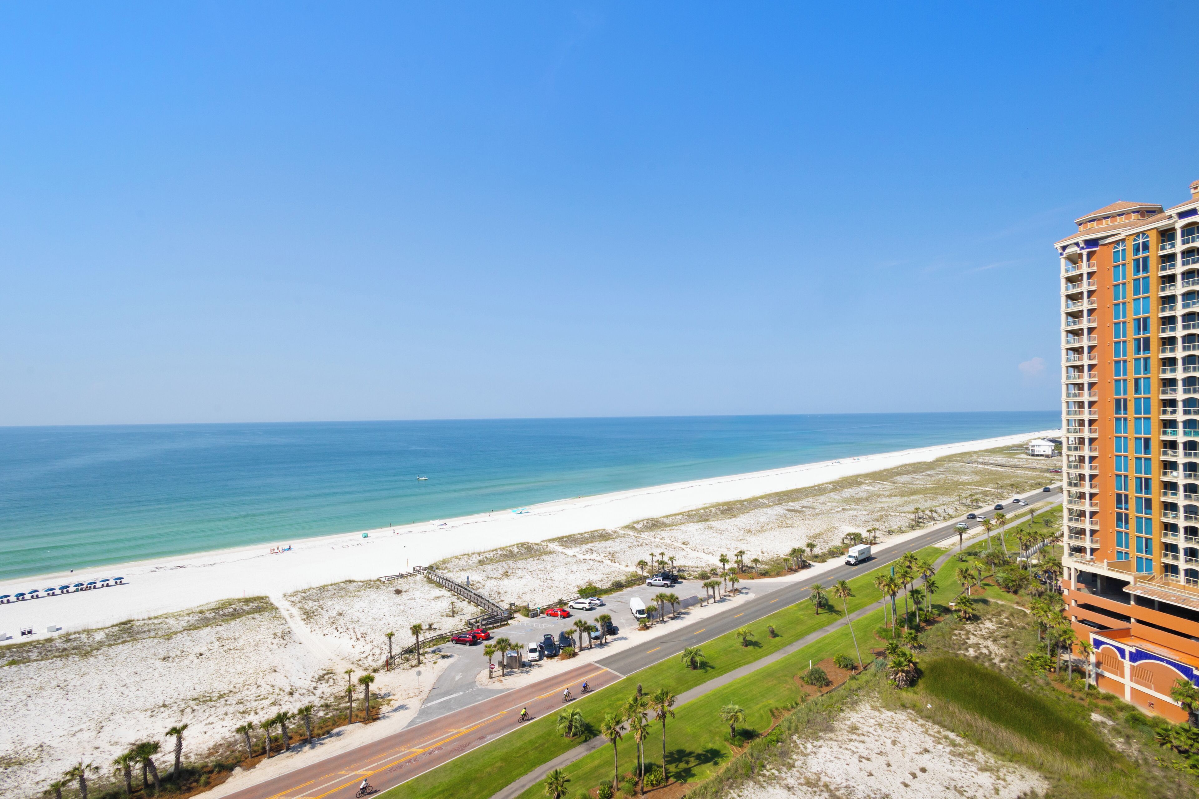 A beachfront hotel with a road leading to the beach, a parking area, and a view of the ocean.