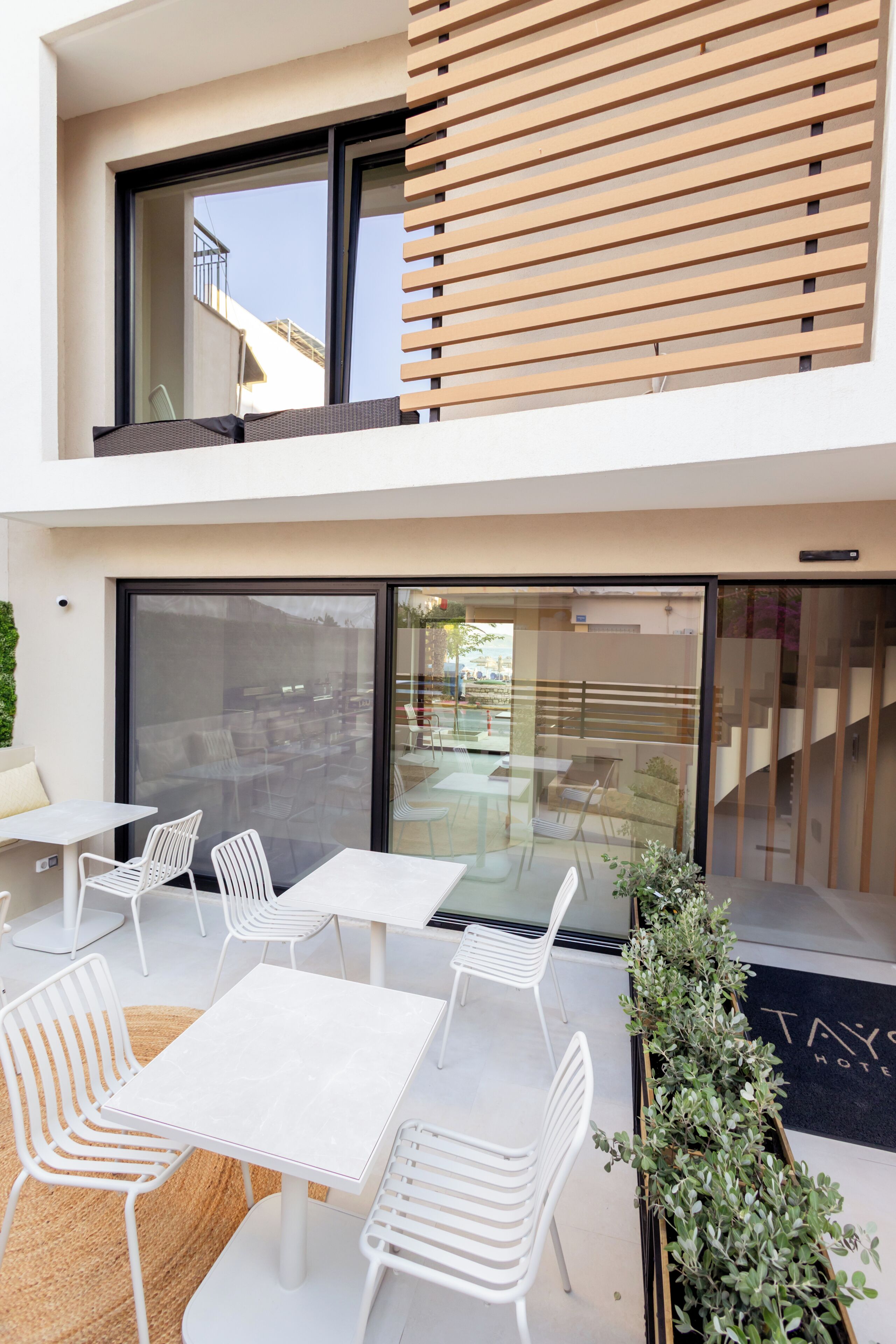 Un espace extérieur moderne, aménagé avec des tables et des chaises blanches, une porte vitrée donnant sur un balcon et un mur en bois à lattes.