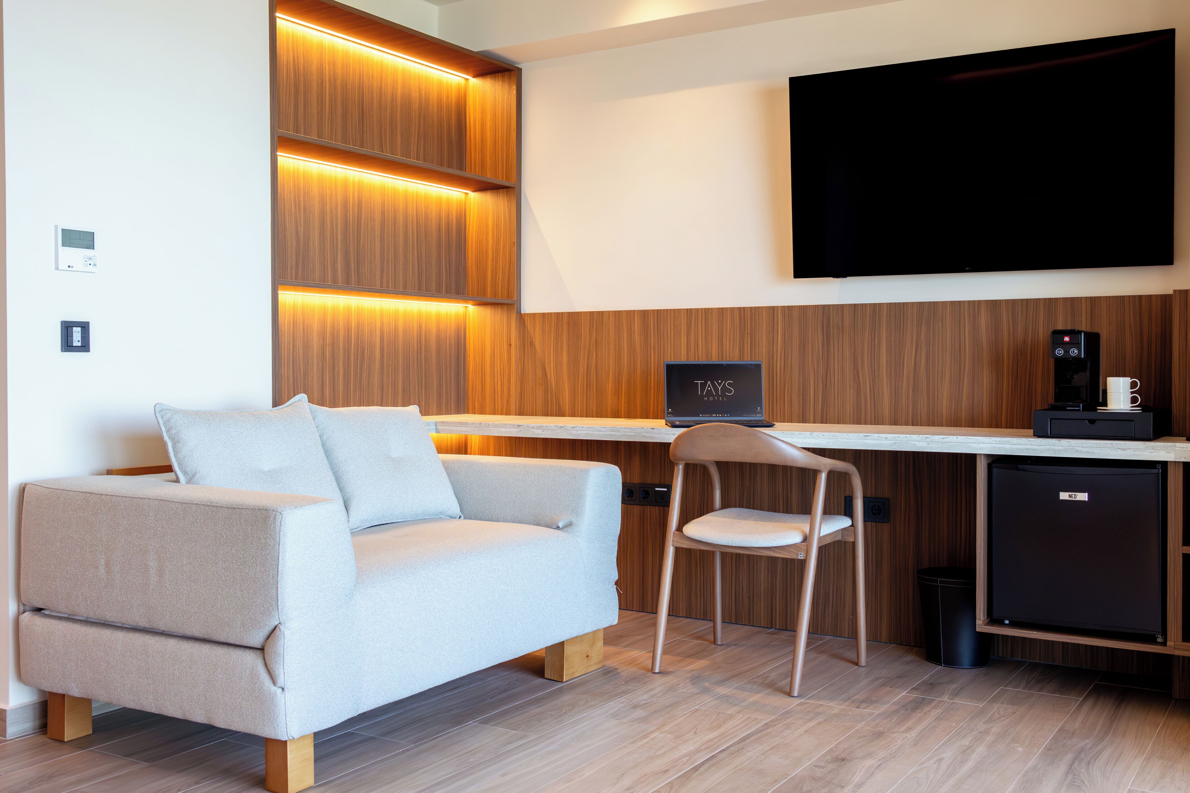 Una habitación de hotel moderna con sofá, escritorio, silla, televisor, cafetera y minibar.