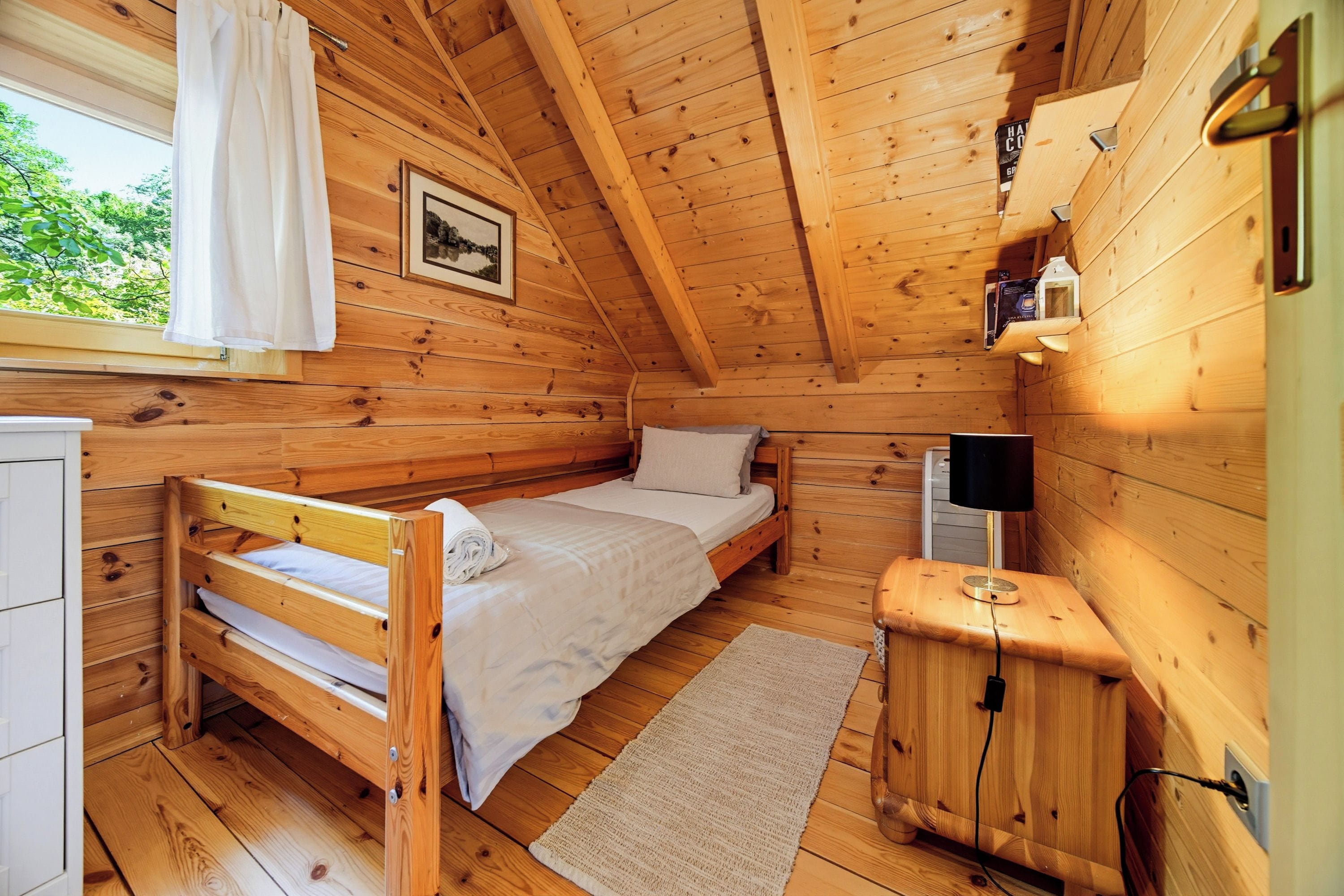 A wooden cabin bedroom with a bunk bed, a bedside table with a lamp, a framed picture on the wall, and a window with curtains.