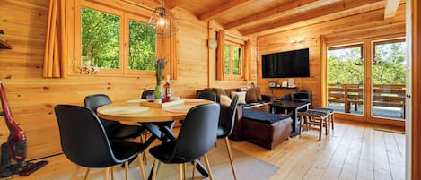 A wooden cabin interior with a dining area, seating, a TV, and large windows.
