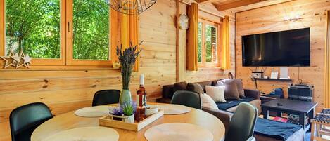 A wooden cabin interior with a dining table, chairs, a sofa, a TV, and large windows.