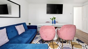 Ein modernes Wohnzimmer mit einem blauen Sofa, rosafarbenen Sesseln, einem weißen Tisch und einem an der Wand befestigten Flachbildfernseher.