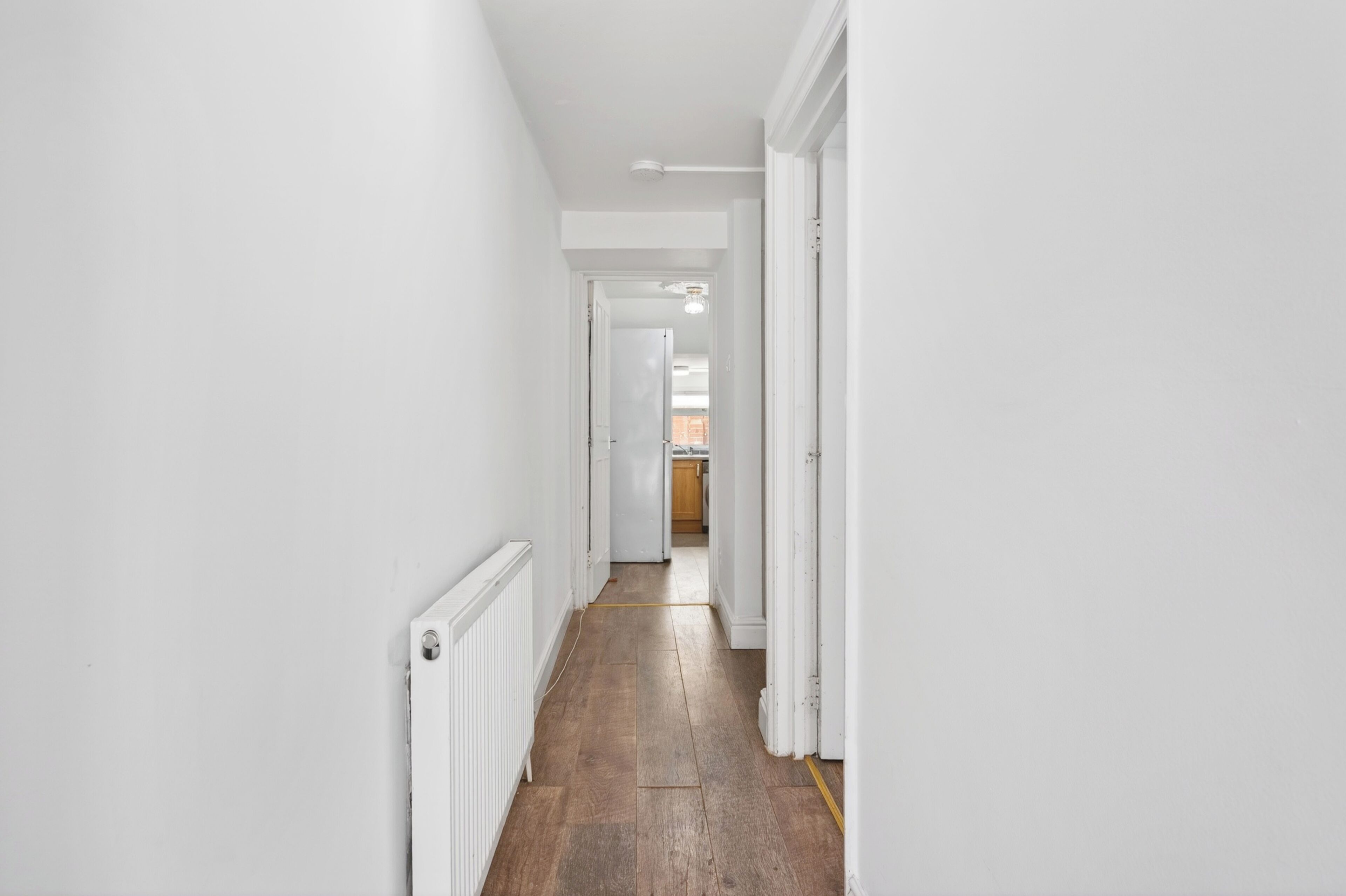 A narrow hallway with white walls and wooden flooring, leading to a kitchen area.