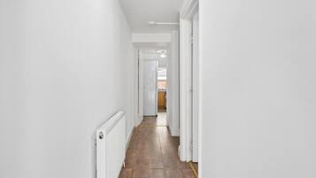 A narrow hallway with white walls and wooden flooring, leading to a kitchen area.