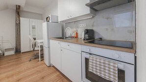 Une cuisine compacte avec des meubles blancs, un four encastré, une machine à café et un petit coin repas avec une chaise.