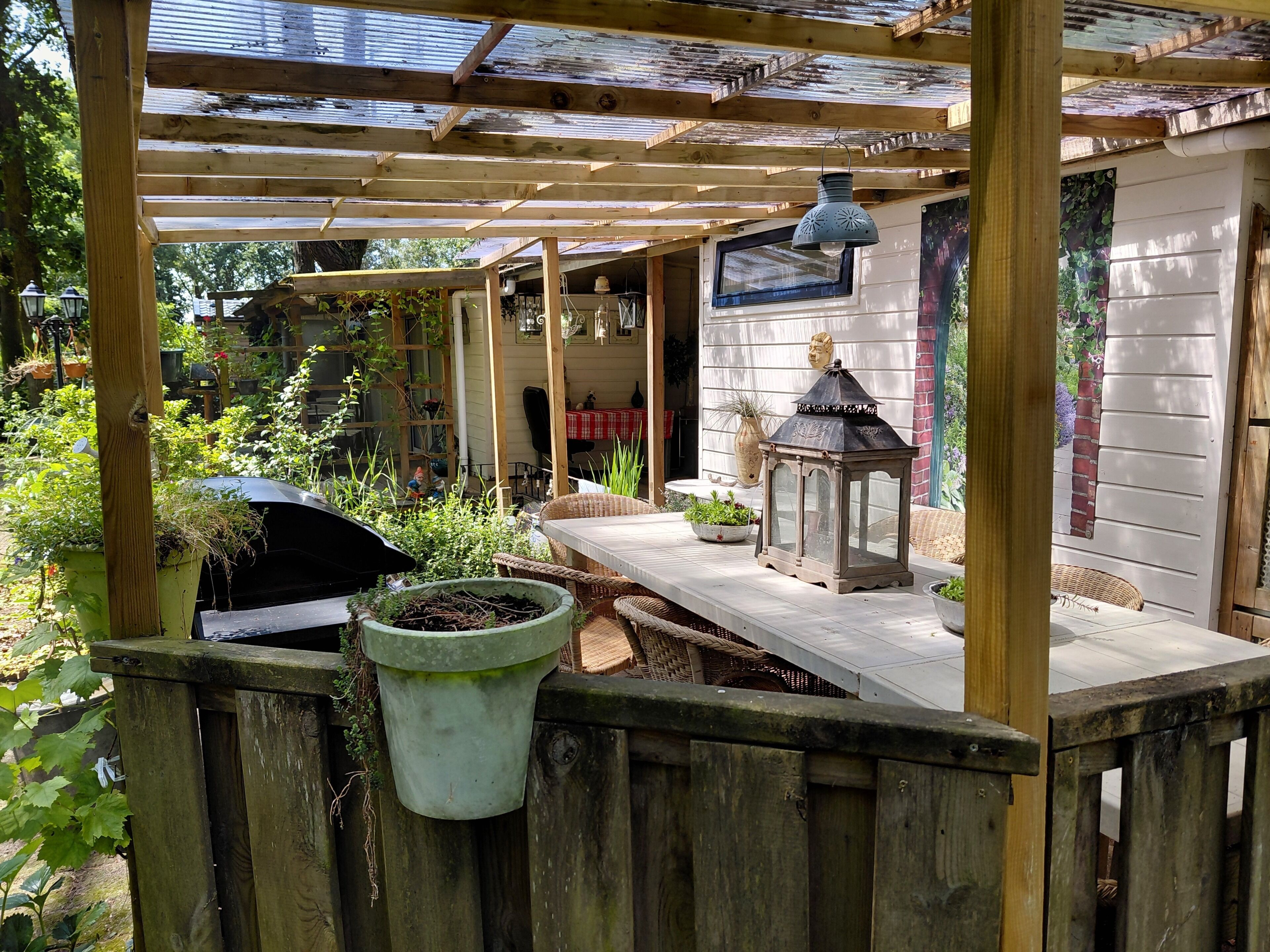 A wooden pergola with a table and lanterns, surrounded by potted plants and a brick wall.