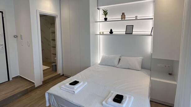 Una habitación de hotel moderna con una cama grande, una mesita de noche, un estante empotrado con objetos decorativos y una puerta que da a un baño.