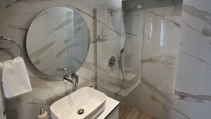 Un baño moderno con tocador de mármol, un espejo redondo, cabina de ducha de vidrio y suelo de madera.