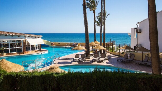 Ein Poolbereich mit Liegestühlen, Palmen und Blick aufs Meer.