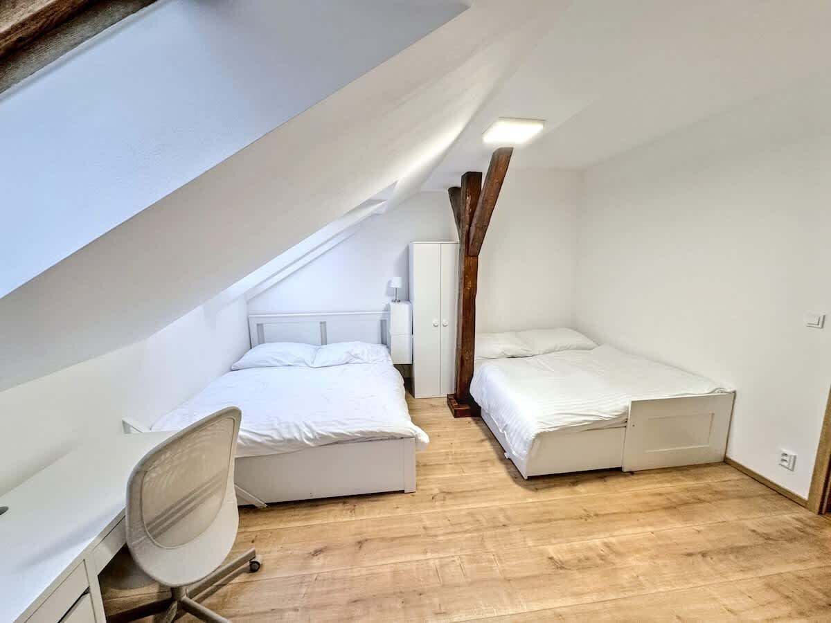 A small, single-room apartment with a sloped ceiling, two beds, a desk, and a chair.