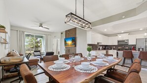 Un coin repas avec une table en bois dressée pour six personnes, entourée de chaises en cuir, d’un ventilateur de plafond et offrant une vue sur la cuisine en arrière-plan.
