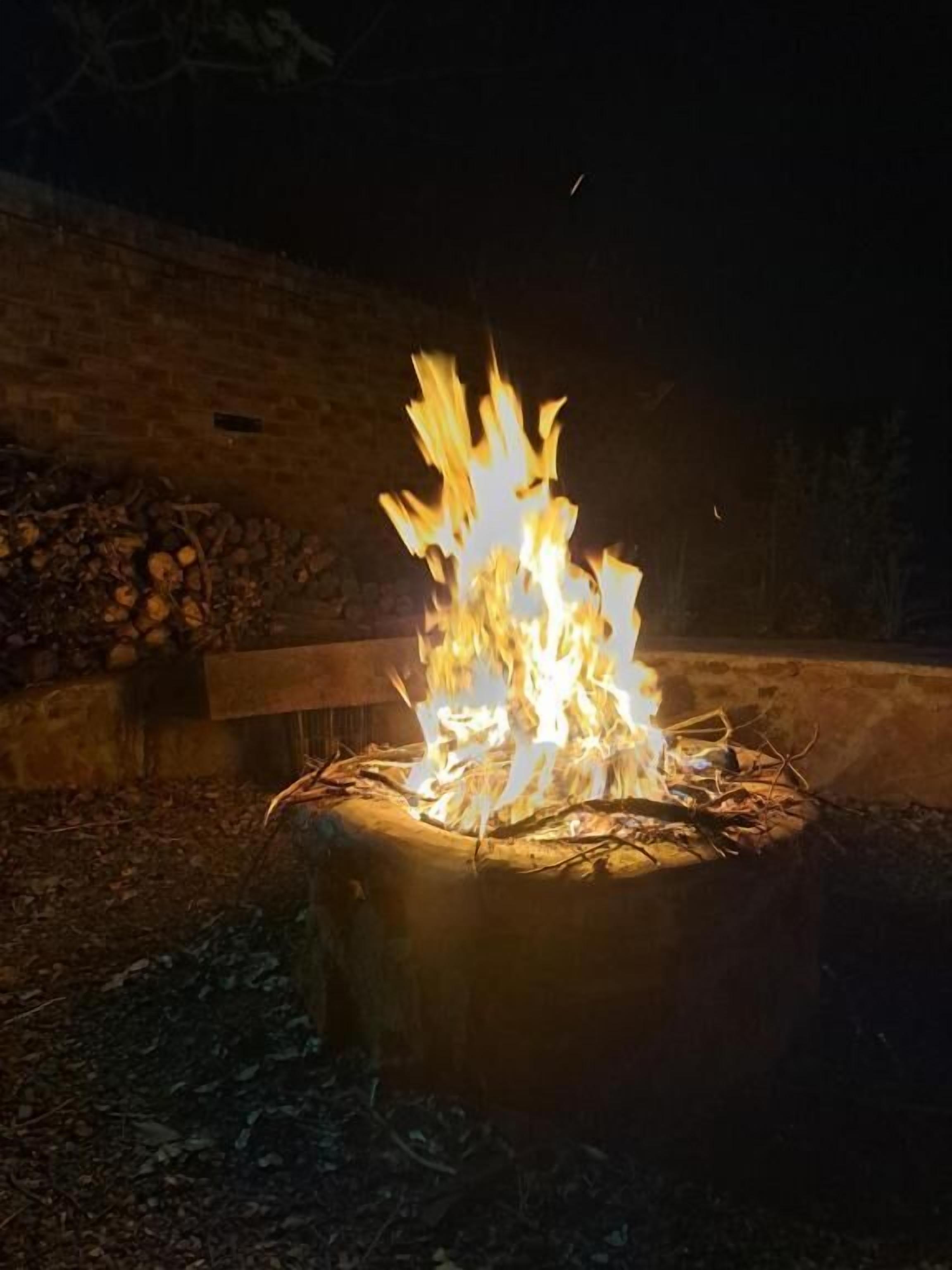 Una fogata en un brasero circular de piedra rodeado por un muro de ladrillo y una pila de leña.