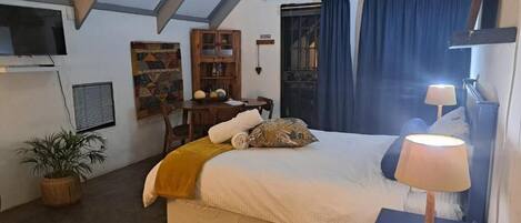 A bedroom with a bed, a bedside table with a lamp, a TV mounted on the wall, a window with curtains, a bookshelf, and a potted plant.