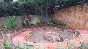 Um jardim circular com uma lareira no centro, rodeado por um canteiro de tijolos e diversas plantas.