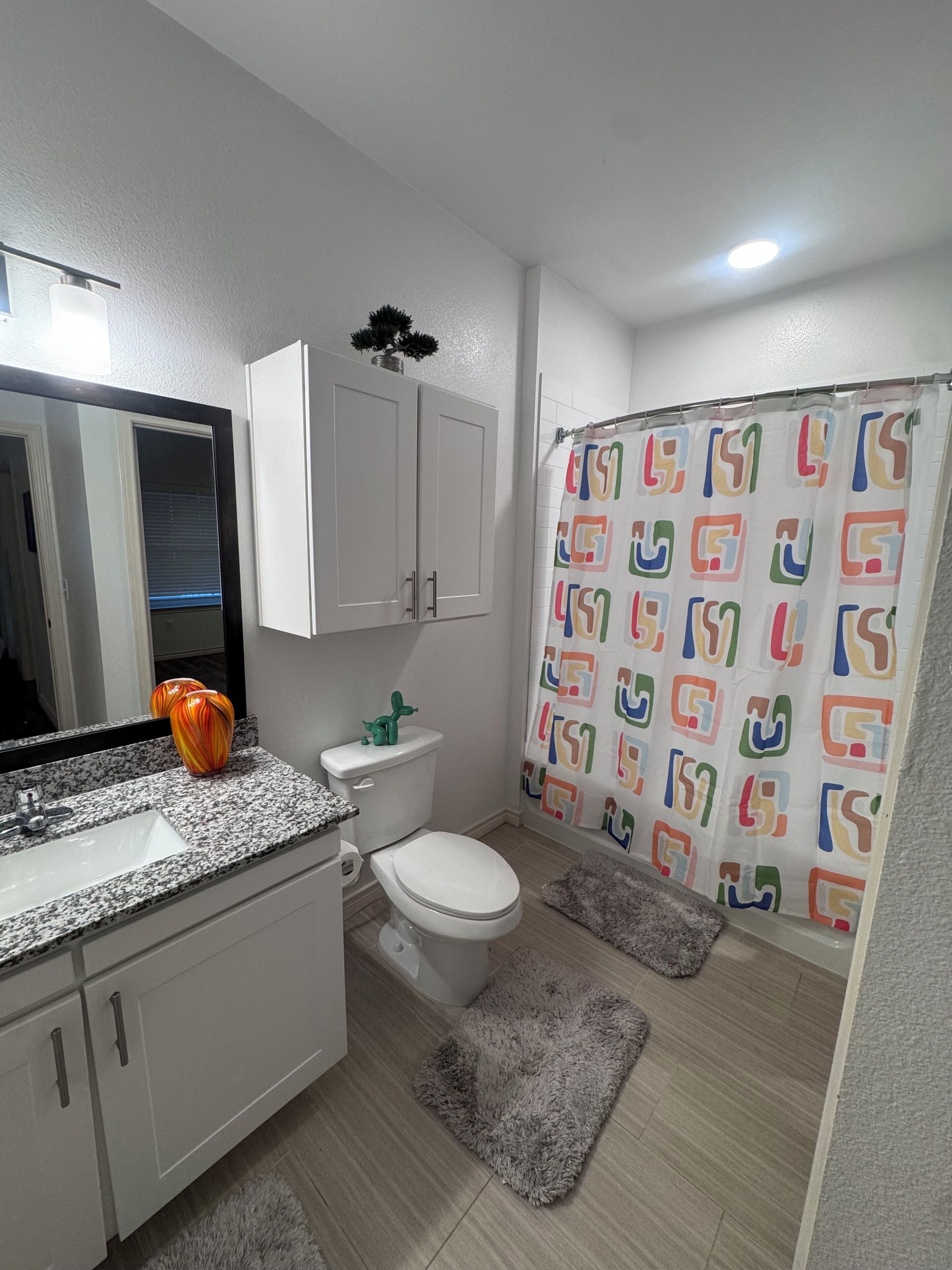 A bathroom with a toilet, sink, mirror, cabinets, shower curtain, and rugs.