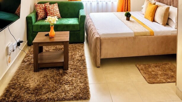 A modern hotel room with a green sofa, a bed with a beige bedspread, a small table with a vase, and a large window with curtains. - Beautiful Place to Unwind – Executive Studio at Garden City Mall, Garden 237 (Nairobi)